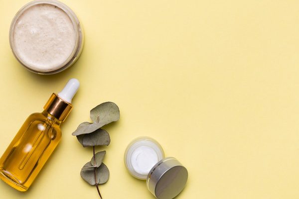 Natural skincare products including a dropper bottle, cream jars, and eucalyptus leaves on a yellow background.