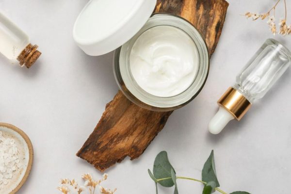 Open jar of cream, dropper bottle, and natural elements like wood and eucalyptus leaves for skincare.